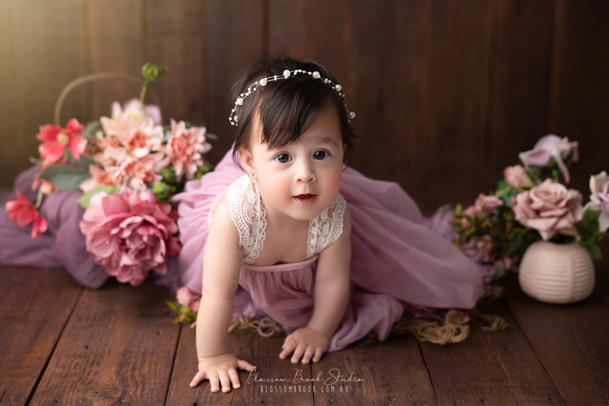 A sweet baby girl in a mauve dress crawling on wooden floor surrounded by pink flowers — Baby Portrait Photography Sydney by Blossom Brook Studio.