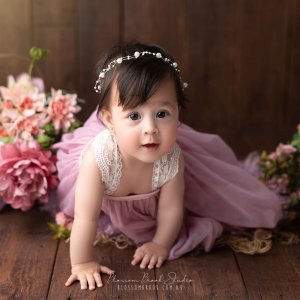 Celine | 20 days | Newborn photography 2 A sweet baby girl in a mauve dress crawling on wooden floor surrounded by pink flowers — Baby Portrait Photography Sydney by Blossom Brook Studio.