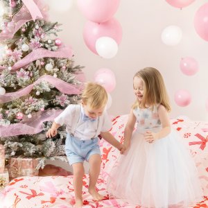 Two children holding hands and playing during a Christmas Family Photoshoot, surrounded by pink decorations and festive styling, captured as Children Christmas Photography in a bright studio.