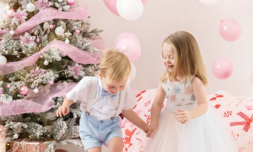 Two children holding hands and playing during a Christmas Family Photoshoot, surrounded by pink decorations and festive styling, captured as Children Christmas Photography in a bright studio.