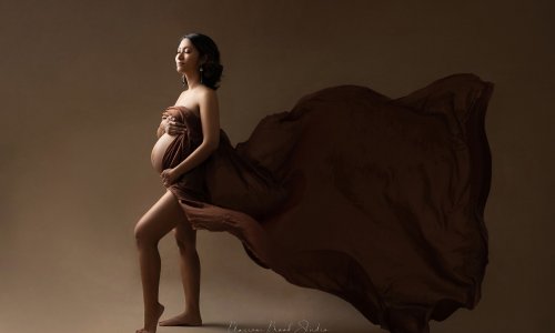 Maternity Portraits of a pregnat lady covered by a piece of brown fabric in the wind, with pleasant expression, holding her belly.