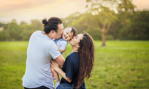 Loving parents kissing their smiling child during an outdoor family photoshoot in a beautiful green park at sunset.