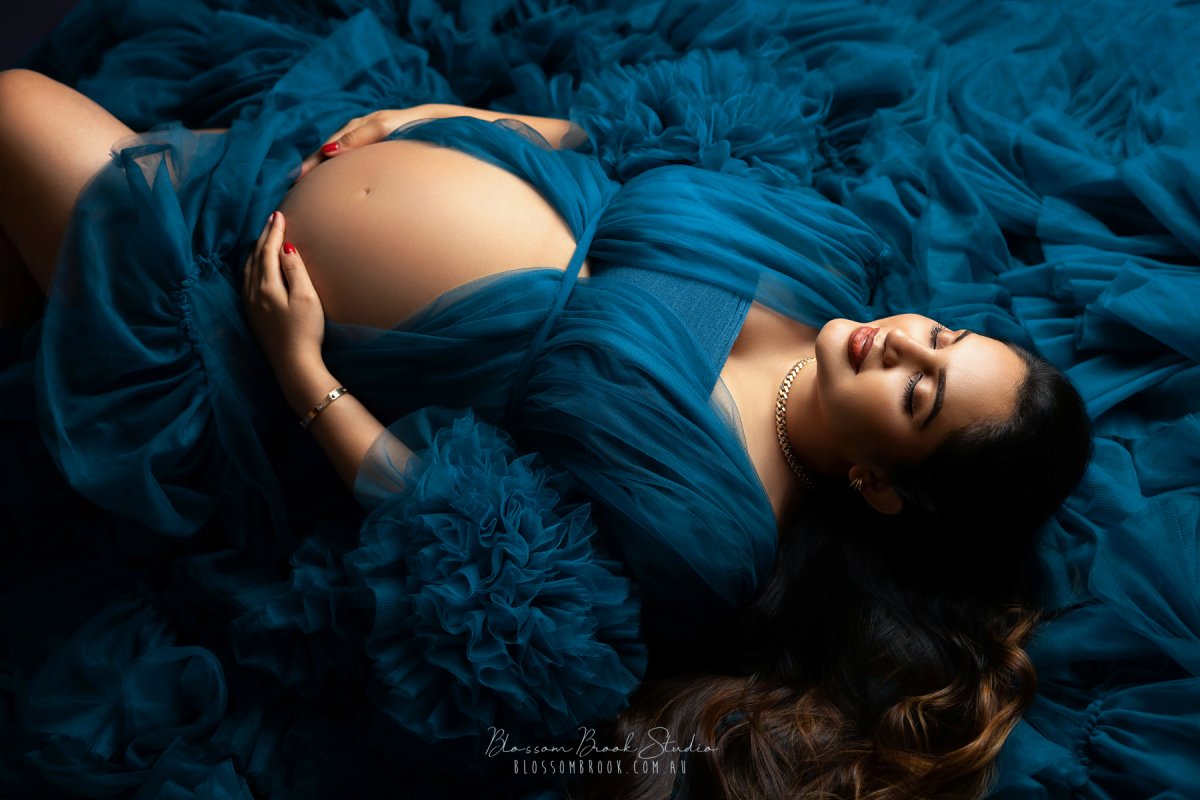 A maternity photoshoot of a couple on a deep blue background. The woman wears a flowing teal gown with ruffles, showcasing her baby bump, while her partner sits beside her, gently holding her belly and kissing her temple. Captured by Blossom Brook Studio.