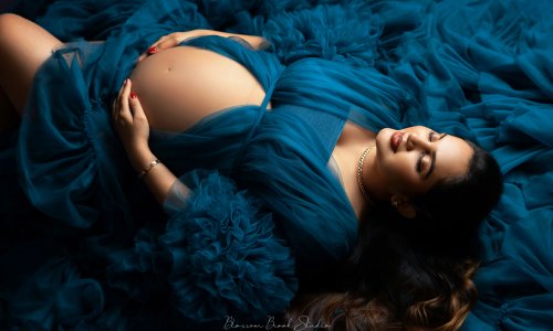 A maternity photoshoot of a couple on a deep blue background. The woman wears a flowing teal gown with ruffles, showcasing her baby bump, while her partner sits beside her, gently holding her belly and kissing her temple. Captured by Blossom Brook Studio.