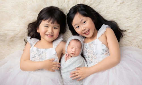 Two older sisters smiling while holding their newborn sibling wrapped in a blanket during a professional newborn photoshoot.