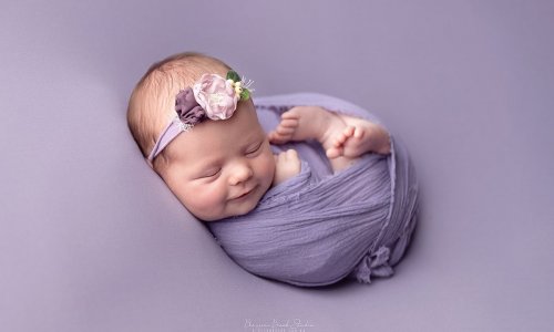Newborn baby girl peacefully swaddled in purple wrap with floral headband during professional newborn photography Sydney session.