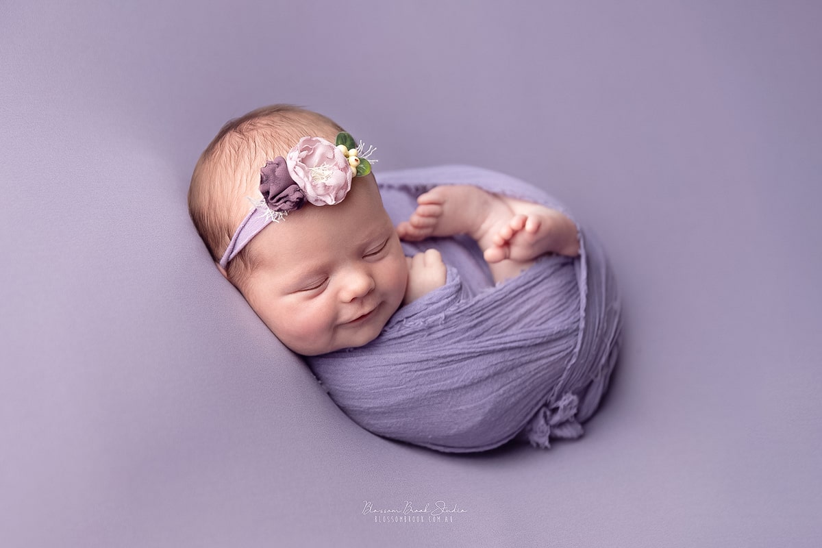 Newborn baby girl peacefully swaddled in purple wrap with floral headband during professional newborn photography Sydney session.